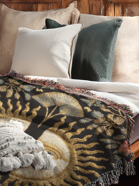 Jacquard-woven cotton blanket featuring twin celestial suns—two serene faces meeting at center—with radiant golden rays on a charcoal ground, bordered by folkloric botanicals and fan blossoms in sage, sky, and garnet. Crafted from 100% soft cotton yarn; the artwork is woven, not printed. Reversible with a color-negative back and finished with a protective 2-inch multicolor fringe. Sizes: 52×37, 50×60, 60×80 for wall, sofa, or queen-bed layering.